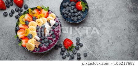Healthy summer acai smoothie bowl with chia seeds, fresh banana, strawberry, blueberry, cocos, kiwi top view, rustic concrete background with spoon. Copy space 106217692
