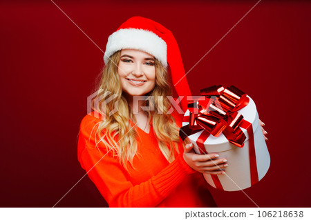 a beautiful young woman in a Santa Claus hat with a gift on a red background.  106218638