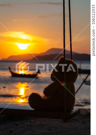 lonely Teddy Bear sitting on a swing in front Rawai beach to see sunrise. scenic reflection of beautiful sunshine above fishing boats are floating in the Rawai sea. beautiful golden sky background. lonely Teddy Bear sitting on a swing in front Rawai beach to see sunrise. scenic reflection of beautiful sunshine above fishing boats are floating in the Rawai sea. beautiful golden sky background. 106222035