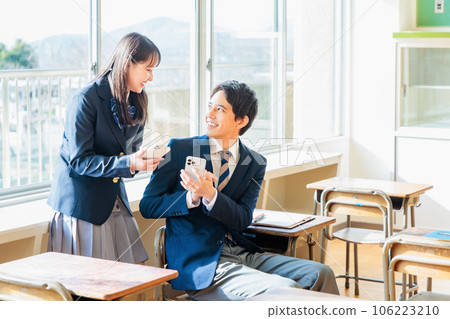 一名高中女生在教室裡一邊看著智能手機一邊與一名男學生交談 106223210