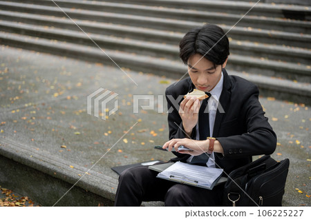 A busy Asian businessman is checking time on his watch and eating a sandwich on a staircase 106225227