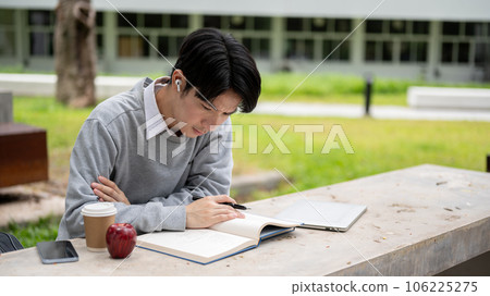 A handsome and smart Asian college man is focusing on reading a book in the campus park 106225275