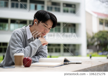 A handsome Asian college man is focusing on reading a book at a table in the campus park 106225276