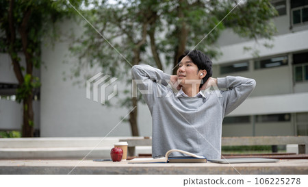 A happy and relaxed Asian male student is stretching his arms while reading books in a campus park 106225278