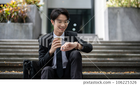 A smart businessman is sipping coffee and checking time on his watch while sitting on a staircase A smart businessman is sipping coffee and checking time on his watch while sitting on a staircase 106225326