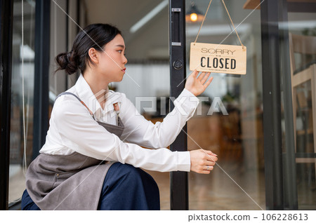 A sad and upset Asian female coffee shop owner is hanging a close sign at the entrance door 106228613