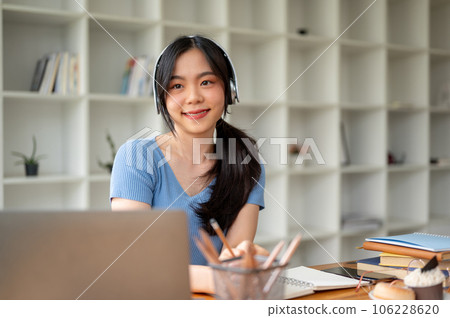 A beautiful Asian female college student smiling, wearing headphones, while sitting at her table 106228620