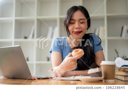 A pretty Asian woman enjoying her snack time, eating doughnuts at her study table at home. 106228645