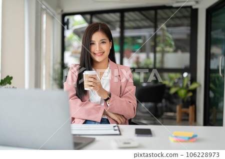 A charming Asian businesswoman smiles at the camera and sits at her desk 106228973