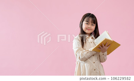 A bright Asian girl is holding a book while standing against an isolated pink studio background. 106229582