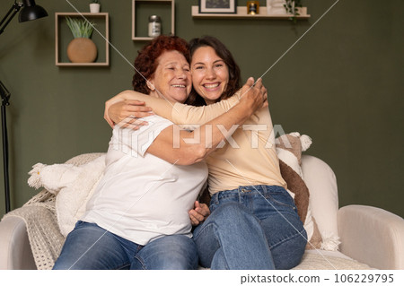 Portrait of a modern happy grandmother and granddaughter sitting in the living room during the day on the sofa, care hugging and looking away Portrait of a modern happy grandmother and granddaughter sitting in the living room during the day on the sofa, care hugging and looking away 106229795