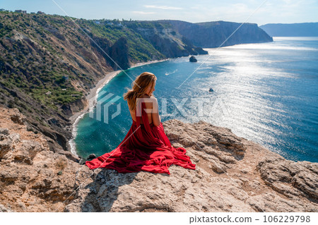 A girl with flowing hair in a long red dress sits on a rock above the sea. The stone can be seen in the sea. 106229798