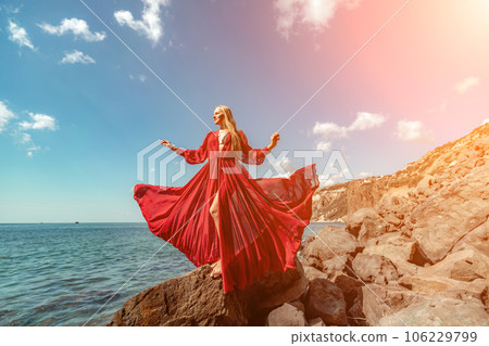 Red dress sea woman. A blonde with flowing hair in a long flowing red dress stands on a rock near the sea. Travel concept, photo session at sea Red dress sea woman. A blonde with flowing hair in a long flowing red dress stands on a rock near the sea. Travel concept, photo session at sea 106229799
