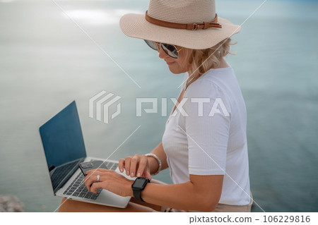 Freelance women sea working on the computer. Good looking middle aged woman typing on a laptop keyboard outdoors with a beautiful sea view. The concept of remote work. 106229816