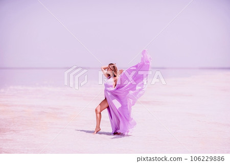Woman pink salt lake. Against the backdrop of a pink salt lake, a woman in a long pink dress takes a leisurely stroll along the white, salty shore, capturing a wanderlust moment. 106229886