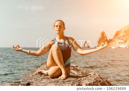 Yoga on the beach. A happy woman meditating in a yoga pose on the beach, surrounded by the ocean and rock mountains, promoting a healthy lifestyle outdoors in nature, and inspiring fitness concept. Yoga on the beach. A happy woman meditating in a yoga pose on the beach, surrounded by the ocean and rock mountains, promoting a healthy lifestyle outdoors in nature, and inspiring fitness concept. 106229899