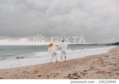 Women sea walk friendship spring. Two girlfriends, redhead and blonde, middle-aged walk along the sandy beach of the sea, dressed in white clothes. Against the backdrop of a cloudy sky and the winter 106229900
