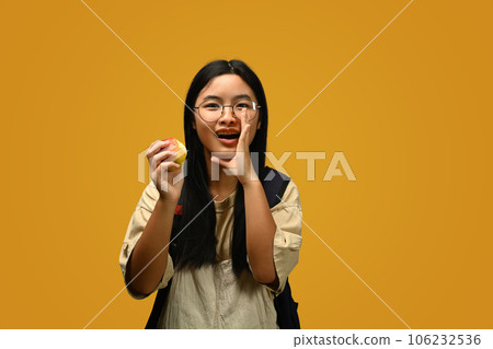 Happy schoolgirl wearing glasses holding an apple on yellow background. Educational, learning and back to school concept 106232536