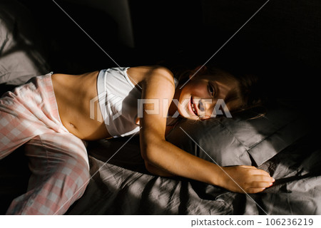 Girl lies on a bed with dark bedding in the sun and smiles in the early morning Girl lies on a bed with dark bedding in the sun and smiles in the early morning 106236219