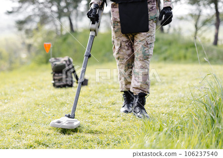 A man in a military uniform and bulletproof vest works in the forest with a metal detector. A minesweeper performs work on demining the territory 106237540