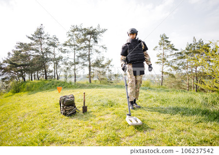 A man in a military uniform and bulletproof vest works in the forest with a metal detector. A minesweeper performs work on demining the territory A man in a military uniform and bulletproof vest works in the forest with a metal detector. A minesweeper performs work on demining the territory 106237542