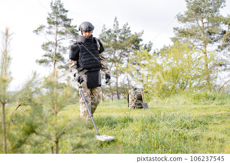 A man in a military uniform and bulletproof vest works in the forest with a metal detector. A minesweeper performs work on demining the territory A man in a military uniform and bulletproof vest works in the forest with a metal detector. A minesweeper performs work on demining the territory 106237545