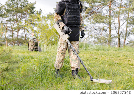 A man in a military uniform and bulletproof vest works in the forest with a metal detector. A minesweeper performs work on demining the territory A man in a military uniform and bulletproof vest works in the forest with a metal detector. A minesweeper performs work on demining the territory 106237548