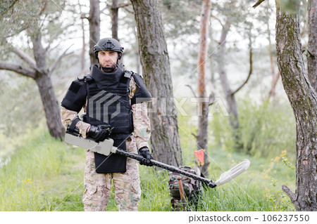 A man in a military uniform and bulletproof vest works in the forest with a metal detector. A minesweeper performs work on demining the territory 106237550