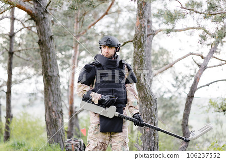 A man in a military uniform and bulletproof vest works in the forest with a metal detector. A minesweeper performs work on demining the territory A man in a military uniform and bulletproof vest works in the forest with a metal detector. A minesweeper performs work on demining the territory 106237552