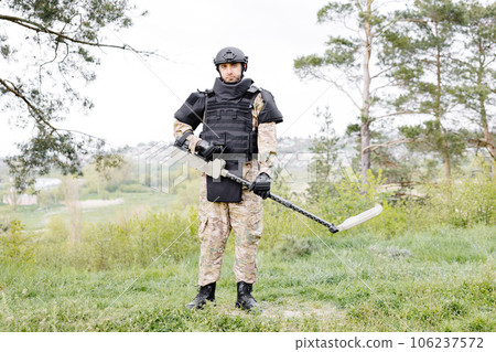 A man in a military uniform and bulletproof vest works in the forest with a metal detector. A minesweeper performs work on demining the territory 106237572