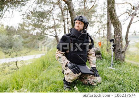 A man in a military uniform and a bulletproof vest works in the forest to demine the territory. A man inspects the area before starting work A man in a military uniform and a bulletproof vest works in the forest to demine the territory. A man inspects the area before starting work 106237574