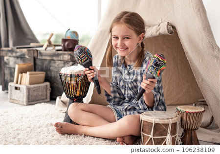 Smiling little girl shaking music maracas sitting in room with djembe drums and wigwam 106238109