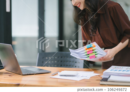 The businesswoman's hands are busy working amidst stacks of paper files, searching and checking for unfinished documents among the folders and papers on her cluttered office desk. The businesswoman's hands are busy working amidst stacks of paper files, searching and checking for unfinished documents among the folders and papers on her cluttered office desk. 106249872