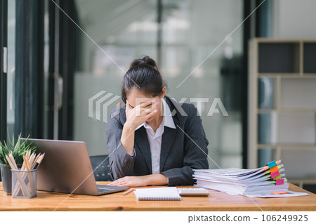 The stressed and exhausted millennial Asian businesswoman is seen sitting at her office desk with her hand on her head, indicating a hard working day where she is overloaded with work. 106249925
