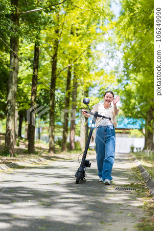 A young and cute woman who visits a beautiful green tree-lined street with an electric kickboard and waves her hand｜Electric kickboard image 106249990