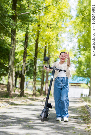 A young and cute woman who visits a beautiful green tree-lined street with an electric kickboard and waves her hand｜Electric kickboard image 106249991