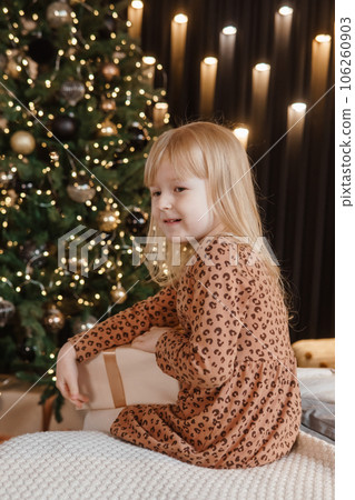 A little blonde girl is sitting on a soft bed in a festive interior decorated in a New Year's style. A child holds a Christmas gift in a craft package. 106260903
