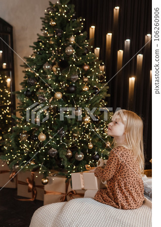 A little blonde girl is sitting on a soft bed in a festive interior decorated in a New Year's style. A child holds a Christmas gift in a craft package. 106260906