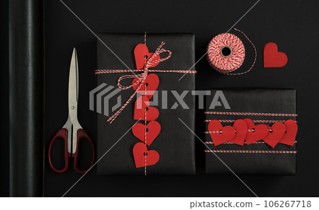 Items for packing gifts for Valentine's Day. Black packing paper, red paper hearts, rope, scissors. Black background. Items for packing gifts for Valentine's Day. Black packing paper, red paper hearts, rope, scissors. Black background. 106267718