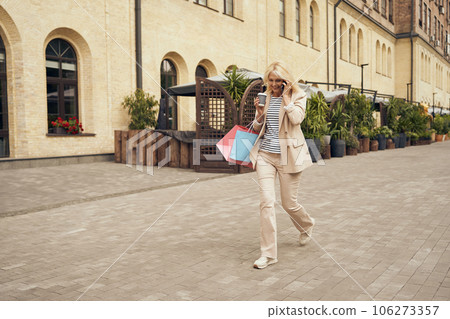 Mature woman walking in city street with coffee and talking phone after shopping. Seasonal sale 106273357
