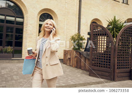 Stylish mature woman with shopping bags walking in city center and talking phone. Seasonal sales 106273358