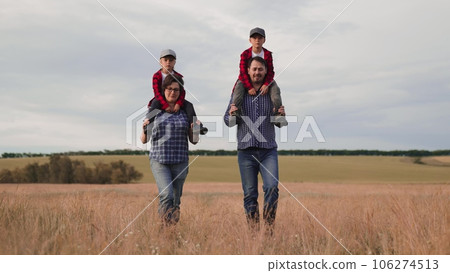 Mother and father farmers carry little boys on shoulders walking together in country field. Farmers parents spend time with cute sons walking across farmland. Farmers with little boys in dry field 106274513