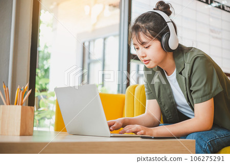 teenage woman wearing headphones sits on her phone while listening to music, playing games, ordering online on the sofa comfortably in a cafe. teenage woman wearing headphones sits on her phone while listening to music, playing games, ordering online on the sofa comfortably in a cafe. 106275291