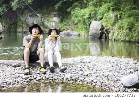 Young man and woman eating grilled corn while admiring the view on the riverbed Young man and woman eating grilled corn while admiring the view on the riverbed 106277732