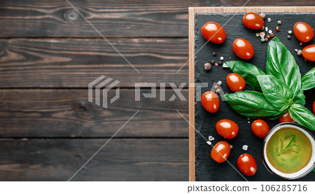 Olive oil and cherry tomatoes with basil leaves, salt and pepper, layout on a black stone board. Ingredients for making Caprese salad. Copy space on a dark wooden background, top view 106285716