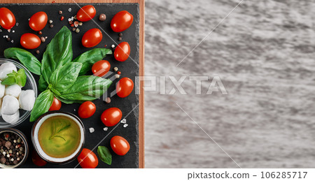 Mozzarella and cherry tomatoes with basil leaves, salt and pepper, layout on a black stone board. Ingredients for making Caprese salad. Top view with copy space for text 106285717