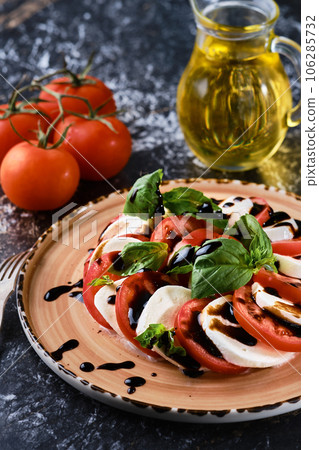 Italian Caprese salad with tomatoes, mozzarella cheese, basil, balsamic vinegar and olive oil. On a ceramic plate. Close-up, selective focus of marble dark table. Vegetarian dish Italian Caprese salad with tomatoes, mozzarella cheese, basil, balsamic vinegar and olive oil. On a ceramic plate. Close-up, selective focus of marble dark table. Vegetarian dish 106285732