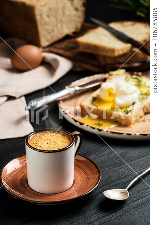 Close-up of cup of espresso coffee with rising steam on black wooden table. On blurred background, soft-boiled egg (poached) in slice of bread, with butter cream and herbs. Breakfast idea Close-up of cup of espresso coffee with rising steam on black wooden table. On blurred background, soft-boiled egg (poached) in slice of bread, with butter cream and herbs. Breakfast idea 106285885