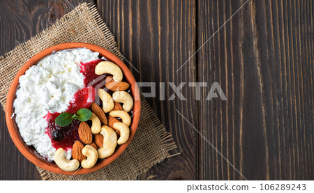 Crumbly fresh cottage cheese with sour cream, cranberry jam and nuts in a traditional clay bowl on a wooden background. Healthy breakfast made from curd, top view with copy space Crumbly fresh cottage cheese with sour cream, cranberry jam and nuts in a traditional clay bowl on a wooden background. Healthy breakfast made from curd, top view with copy space 106289243