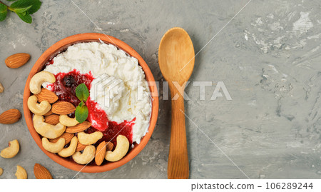 Crumbly fresh cottage cheese with sour cream, cranberry jam and nuts in traditional clay bowl, next to wooden spoon. Neutral gray background. Healthy breakfast made from curd, top view with copy space Crumbly fresh cottage cheese with sour cream, cranberry jam and nuts in traditional clay bowl, next to wooden spoon. Neutral gray background. Healthy breakfast made from curd, top view with copy space 106289244